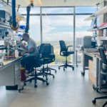 Annapurna Labs: A man looks through a microscope in a cluttered lab space, chairs are haphazardly set through the room, with counter space a premium, a polished off-white floor leads to large windows, which display the Austin skyline that is blue filled with clouds.