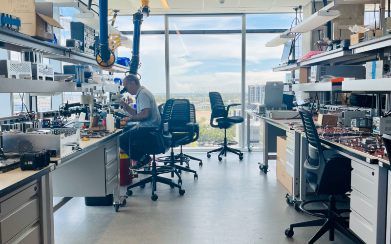 Annapurna Labs: A man looks through a microscope in a cluttered lab space, chairs are haphazardly set through the room, with counter space a premium, a polished off-white floor leads to large windows, which display the Austin skyline that is blue filled with clouds.